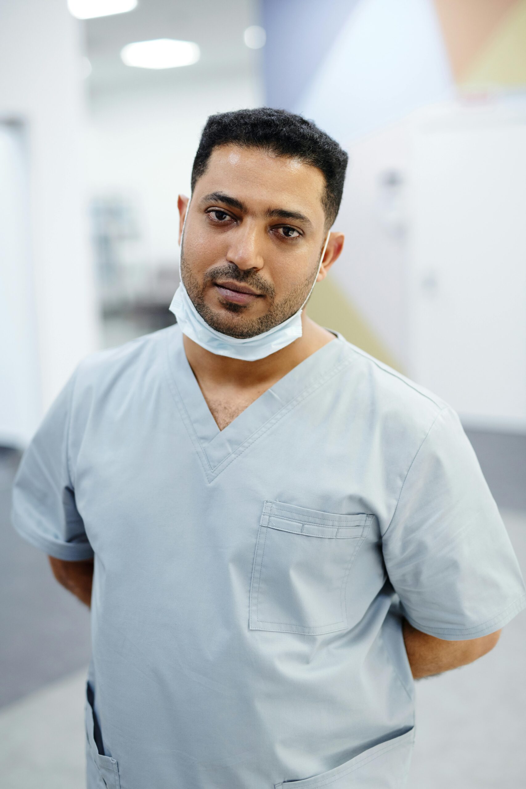 A healthcare worker in blue scrubs standing indoors, with a mask around the neck.